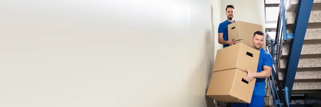 שני מובילים בחולצות כחולות נושאים שלושה קרטונים בחדר מדרגות עם מעקה כחול