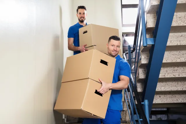 שני סבלים בסרבלים כחולים נושאים קרטונים בחדר מדרגות צר עם מעקה כחול