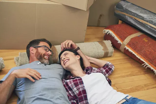 זוג שוכב על רצפת פרקט ליד שטיח מגולגל וקרטונים וצוחק