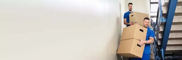 שני מובילים בחולצות כחולות נושאים שלושה קרטונים בחדר מדרגות עם מעקה כחול