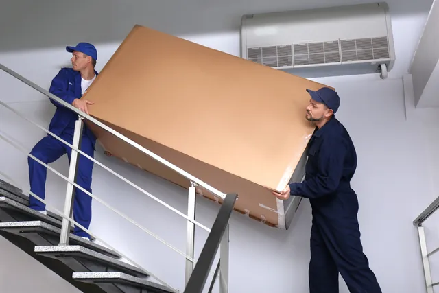 שני מובילים בסרבלים כחולים מעלים במדרגות צרות פריט גדול עטוף בקרטון לצד מעקה מתכת