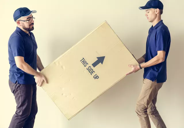 שני מובילים בחולצות כחולות נושאים קרטון שטוח גדול עם חץ שמסמן איזה צד כלפי מעלה