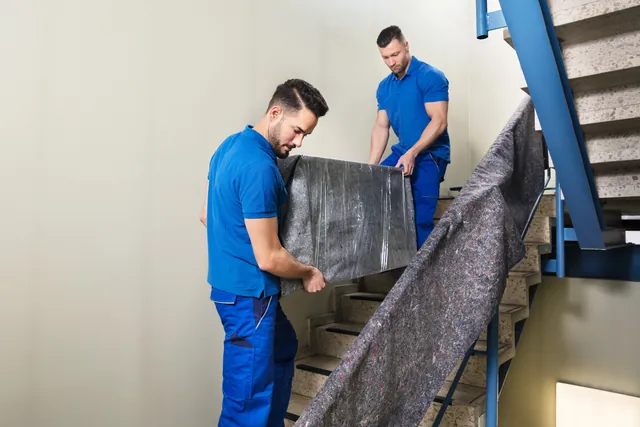 שני מובילים מעבירים רהיט עטוף בשמיכות הגנה בחדר מדרגות עם מעקה כחול — המסלול שאחרי המדרכה
