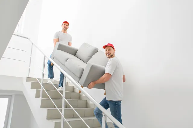 שני מובילים בחולצות לבנות וכובעים אדומים נושאים ספה אפורה במדרגות בניין, לצד מעקה מתכת