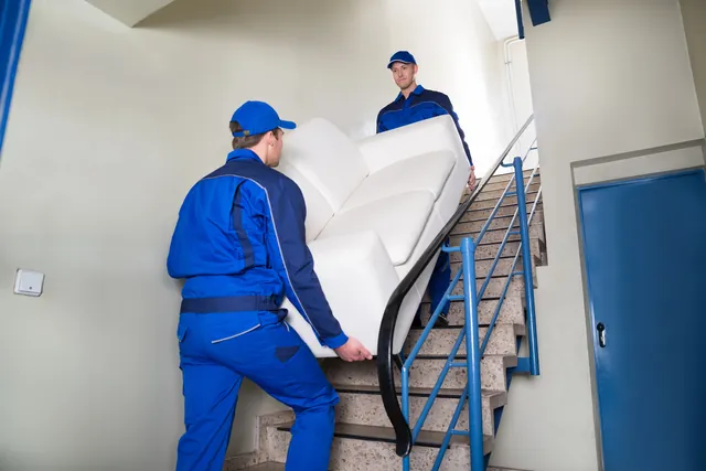 שני מובילים מעלים ספה לבנה בחדר מדרגות צר עם מעקה, ליד הפנייה אל הפודסט