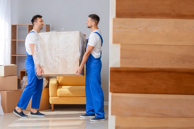 שני מובילים בסרבלים כחולים נושאים פריט גדול עטוף בניילון ליד כורסה צהובה