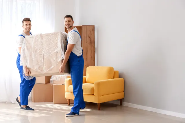 שני מובילים בסרבלים כחולים נושאים רהיט עטוף בניילון, ליד כורסה צהובה