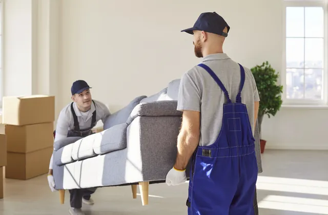 שני מובילים נושאים ספה אפורה בחדר בהיר עם חלון וערימת קרטונים בצד
