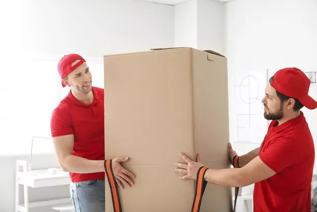 שני מובילים בחולצות אדומות נושאים ברצועות קרטון גבוה וצר של רהיט ארוז